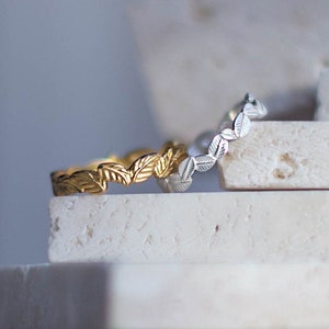 K&ouml;nnte beinhalten: Zwei zarte Ringe mit einem Blattdesign, einer in Gold und einer in Silber, werden auf einem Stapel wei&szlig;er Steinplatten pr&auml;sentiert.