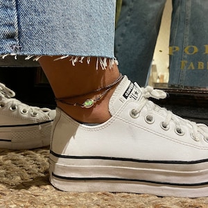 May include: A brown ankle bracelet with a green gemstone charm. The bracelet is worn on a person's ankle with white sneakers.