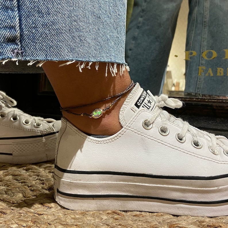 May include: A brown ankle bracelet with a green gemstone charm. The bracelet is worn on a person's ankle with white sneakers.