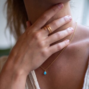 May include: A gold necklace with a teardrop-shaped blue opal pendant. The necklace is worn by a person with a gold ring on their finger.