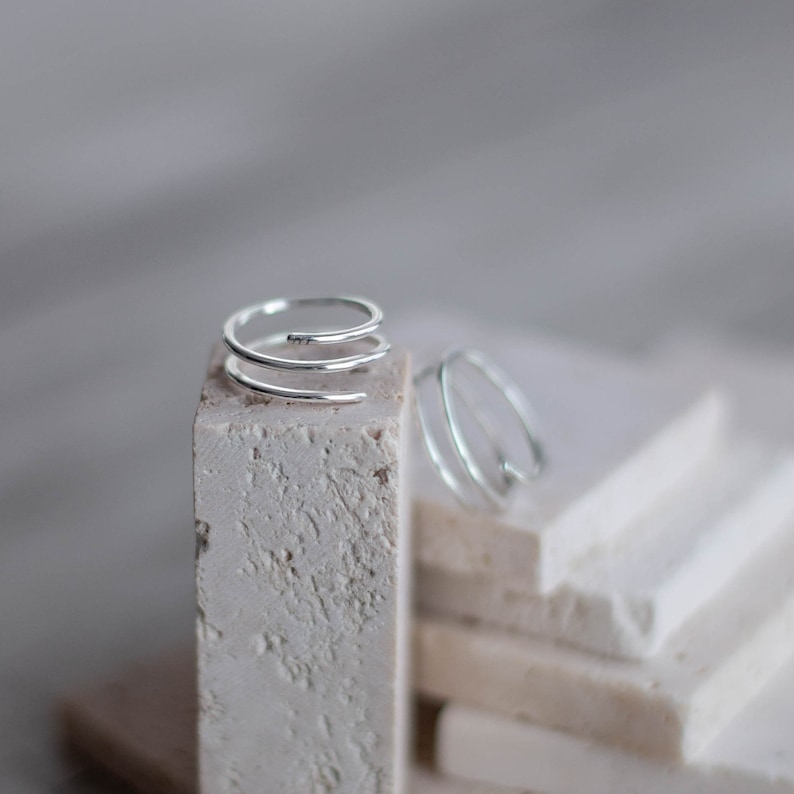 May include: Two silver rings on a white textured surface. The ring on the left is a triple band spiral design. The ring on the right is a single band spiral design.