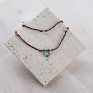 May include: A brown cord necklace with a single teardrop-shaped iridescent blue gemstone pendant and gold beads.