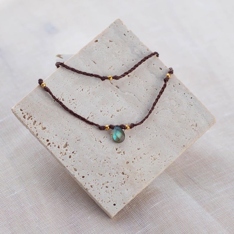May include: A brown cord necklace with a single teardrop-shaped iridescent blue gemstone pendant and gold beads.
