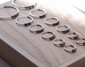 May include: A collection of silver hoop earrings arranged in varying sizes. Each hoop features a thin, circular wire design with a small, textured, decorative metal bead or segment near the closure, showcasing a polished metallic finish.