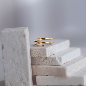 May include: A gold-toned, triple-spiral ring sits on a stack of white rectangular tiles.