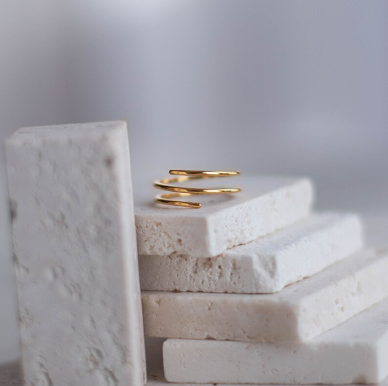 May include: A gold-toned, triple-spiral ring sits on a stack of white rectangular tiles.