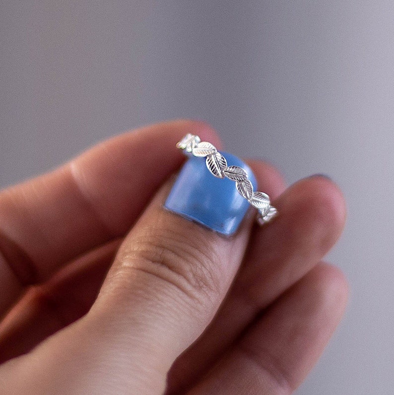 May include: A silver ring with a delicate leaf design. The ring is shown on a finger with blue nail polish.