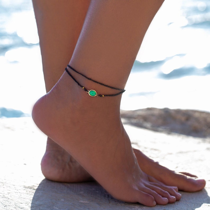 May include: A black ankle bracelet with a green gemstone charm. The bracelet is made of thin cord and has a simple design.