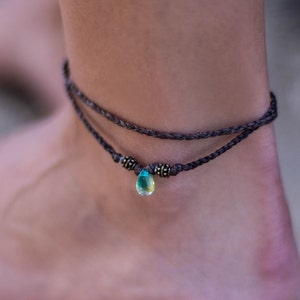 Puede incluir: Un brazalete de tobillo trenzado marrón con un colgante en forma de lágrima de piedra preciosa verde y azul.