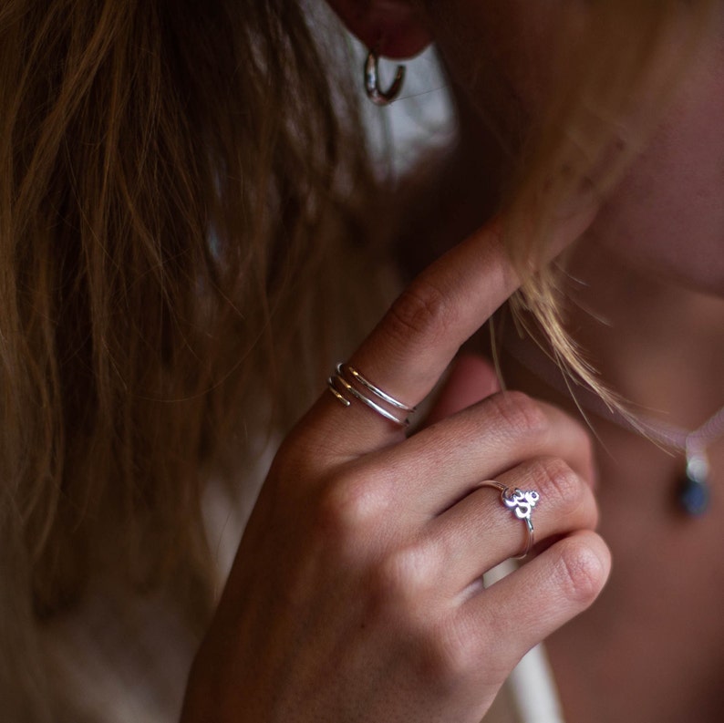May include: A silver ring with a small, abstract design. The ring is worn on the index finger of a person's hand.