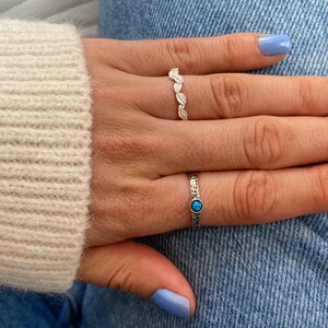 May include: Two silver rings on a hand. One ring has a row of small leaf designs. The other ring has a single blue gemstone.