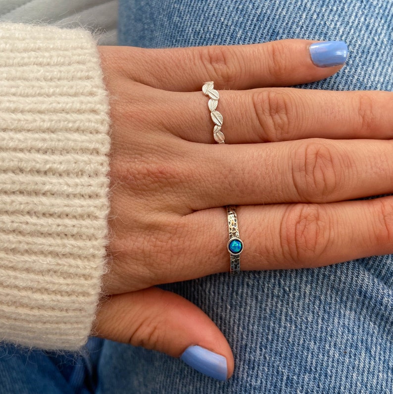 May include: Two silver rings on a hand. One ring has a row of small leaf designs. The other ring has a single blue gemstone.