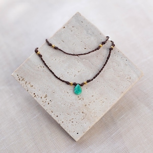 May include: A delicate double-strand necklace with a turquoise teardrop pendant and gold beads. The necklace is made of brown cord.