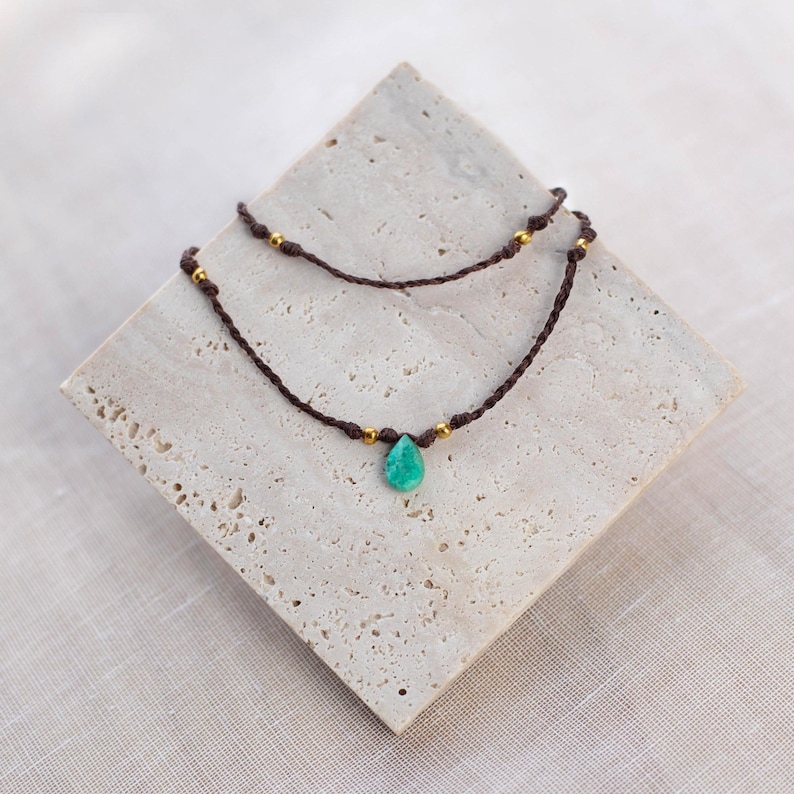 May include: A delicate double-strand necklace with a turquoise teardrop pendant and gold beads. The necklace is made of brown cord.