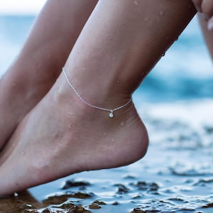 Peut inclure: Un bracelet de cheville en argent avec un petit charme rond en pierre transparente. Le bracelet de cheville est porté à la cheville d'une personne, et le charme en pierre pend.