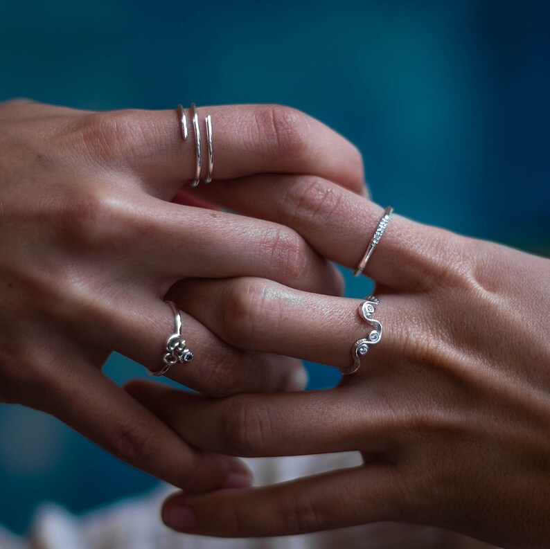 May include: Close-up of two hands wearing silver rings. The rings are delicate and feature various designs, including a simple band, a wavy design, and a ring with a small symbol.