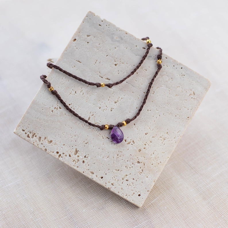 May include: A delicate layered necklace with a purple amethyst pendant and gold beads on brown cord.