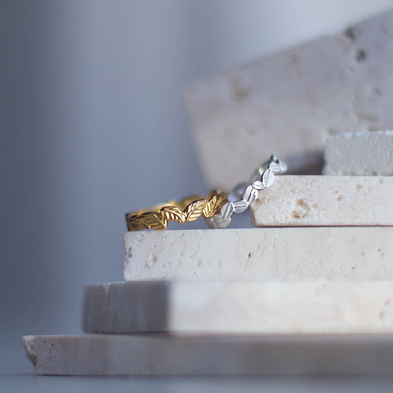 K&ouml;nnte beinhalten: Zwei zarte Ringe mit einem Blattdesign, einer in Gold und einer in Silber, werden auf einem Stapel wei&szlig;er Steinplatten pr&auml;sentiert.