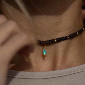 May include: A dark brown woven cord choker necklace featuring small gold-toned beads spaced along the band and a central marquise-shaped pendant with a shimmering iridescent opal stone set in a gold-toned frame with a small decorative bead detail.