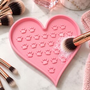 Op de afbeelding: Een hartvormige roze siliconen make-up kwastenreinigingsmat met verhoogde pootafdruktexturen. De mat wordt gebruikt met een make-upkwast met roségouden accenten en witte haren, waarbij tijdens het reinigen zeepsop wordt gevormd.