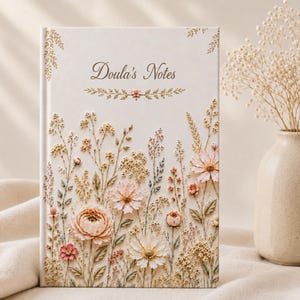 May include: A cream-colored notebook titled "Doula's Notes" with embossed floral designs in soft pink, peach, and beige. The cover features a variety of flowers and foliage, creating a textured, three-dimensional effect. A vase with dried flowers is in the background.