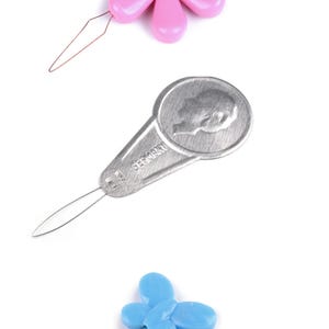 Puede incluir: Tres enhebradores de agujas: uno con mango de plástico rosa en forma de flor, uno con mango de metal plateado grabado con un perfil y la palabra GERMANY, y uno con mango de plástico azul claro en forma de mariposa, cada uno con un bucle de alambre.