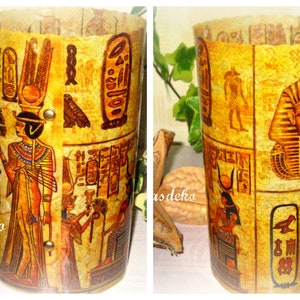 Könnte beinhalten: Zylindrischer dekorativer Behälter mit ägyptischem Thema. Der Behälter ist in einem pergamentartigen Material verpackt, das Hieroglyphen und Illustrationen von Pharaonen und anderen Figuren in Gold-, Braun- und Rottönen zeigt. Der Behälter wird von messingfarbenen Nieten zusammengehalten.