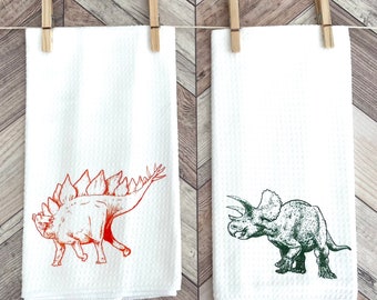 Könnte beinhalten: Zwei weiße Küchentücher mit Dinosaurier-Illustrationen. Das Handtuch links zeigt einen roten Stegosaurus, während das Handtuch rechts einen grünen Triceratops zeigt.