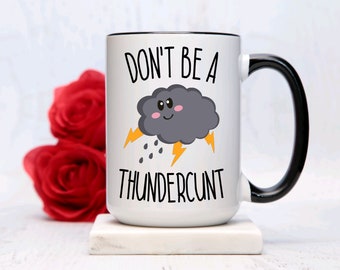Könnte beinhalten: Eine weiße Keramiktasse mit schwarzem Rand und Henkel. Die Tasse zeigt eine schwarz-weiße Grafik einer Cartoon-Gewitterwolke mit Blitzen und Regentropfen. Der Text "Don't be a Thundercunt" ist unter der Wolke gedruckt.