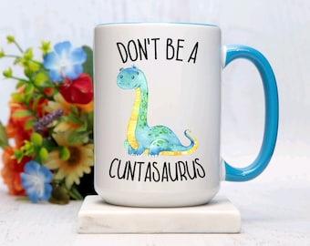 Könnte beinhalten: Weiße Keramiktasse mit blauem Rand und Henkel. Die Tasse zeigt einen Comic-Dinosaurier mit dem Text "Don't be a Cuntasaurus".