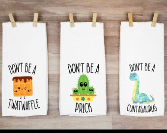 May include: Three white kitchen towels hanging from a string, each with a humorous phrase and illustration. One towel features a waffle with the text "DON'T BE A TWATWAFFLE." Another shows a cactus with the text "DON'T BE A PRICK." The last depicts a dinosaur with the text "DON'T BE A CUNTASAURUS."