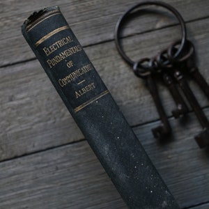 May include: A vintage, dark green book titled "Electrical Fundamentals of Communication" by Albert. The book is lying on a wooden surface with a set of keys in the background.