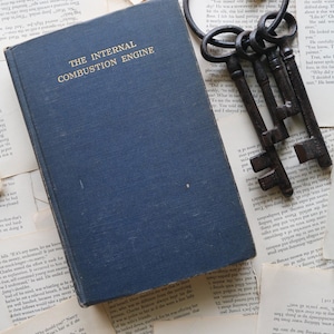 May include: A blue hardback book titled "The Internal Combustion Engine" is lying on a pile of old, white pages. A set of three antique keys with a large ring is on top of the pages.