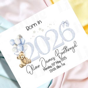 May include: A white card with the text "Born in" above the year "2026". The card features a teddy bear holding blue and silver balloons. The name "Oliver James Boothroyd" is written below the year, along with the date, time, and weight.
