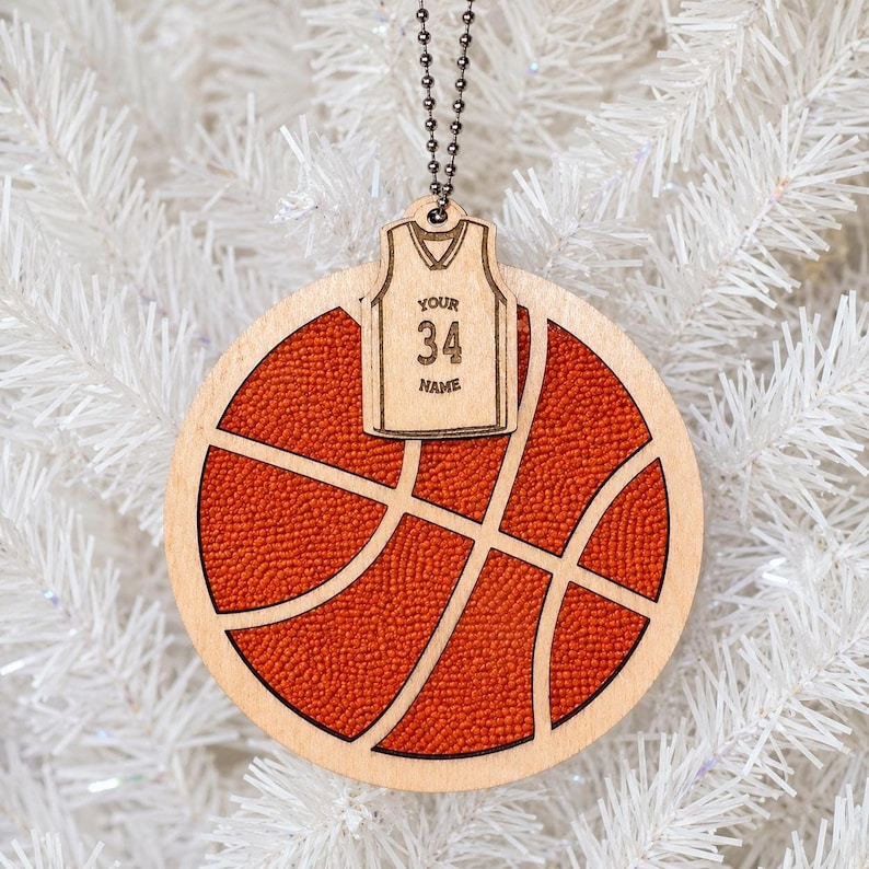 A wooden basketball ornament hangs against a white artificial Christmas tree, with a mini jersey tag attached displaying the text "YOUR 34 NAME".