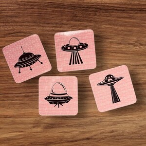 Four square coasters with rounded corners, each featuring a different black UFO illustration on a pink background with a subtle grid pattern, arranged on a wooden surface.