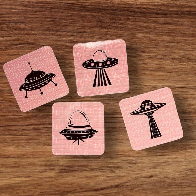 Four square coasters with rounded corners, each featuring a different black UFO illustration on a pink background with a subtle grid pattern, arranged on a wooden surface.