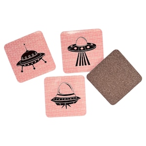 Three square pink coasters with black flying saucer designs, and one coaster turned over to show its brown cork backing, on a white background.