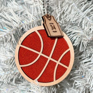 A wooden basketball ornament hangs on a white tree, connected by a chain to a small jersey shaped wood piece that has "YOUR 34 NAME" engraved on it.
