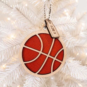 A wooden basketball ornament with a textured orange insert hangs on a lit white Christmas tree, with a small engraved jersey tag attached.