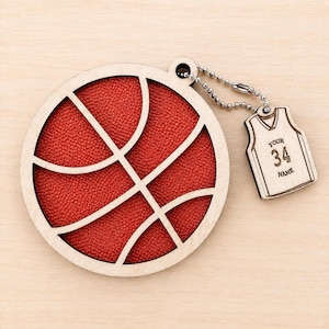 A wooden basketball-shaped ornament rests on a light wood background, attached via a small ball chain to a miniature jersey ornament engraved with "YOUR 34 NAME".