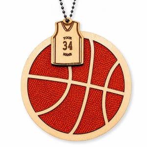 A basketball-shaped wooden ornament with a textured red inlay hangs against a plain white background, joined by a bead chain to a custom jersey plaque.