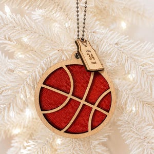 A basketball-shaped wooden ornament with textured orange details hangs in front of a softly lit white artificial Christmas tree.