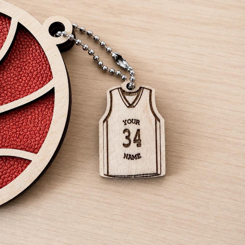 A close-up shot of a wooden basketball ornament with textured red backing and a small ball chain connecting it to an engraved jersey tag reading "YOUR 34 NAME".