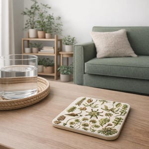 May include: Square coaster with a botanical illustration of various plants in green, brown, and beige. The coaster is on a light wood table, with a glass of water and a woven tray in the foreground. A green sofa and plants are in the background.