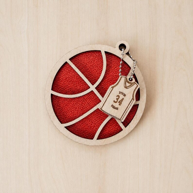 A round wooden basketball-shaped ornament with a red textured background, featuring a small dangling jersey tag engraved with the number 34 and name placeholder.