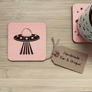 A single square pink coaster with a black flying saucer design, next to a brown tag that reads "Handmade - Fun & Unique!" and a ceramic mug.