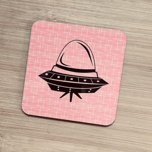 A single square pink coaster with a black flying saucer design featuring a domed top, on a light wood surface.