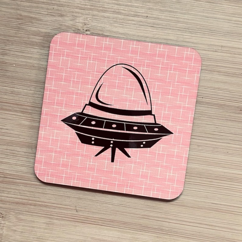 A single square pink coaster with a black flying saucer design featuring a domed top, on a light wood surface.
