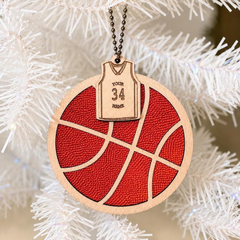 A wooden basketball ornament hangs from a white Christmas tree branch, featuring a textured orange center and an attached mini jersey with the number 34.